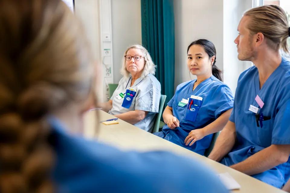 En grupp vårdpersonal som sitter vid ett bord. De tittar mot en fjärde person som inte syns i bild.