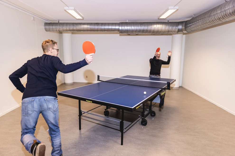 Två personer spelar pingis. De är i en lokal med vita väggar och grått golv. Pingisracken är röda.